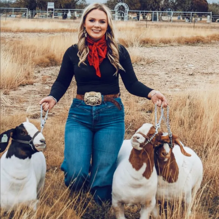 Woman kneels in a dry field, holding two goats on leashes beside her, smiling at the camera.