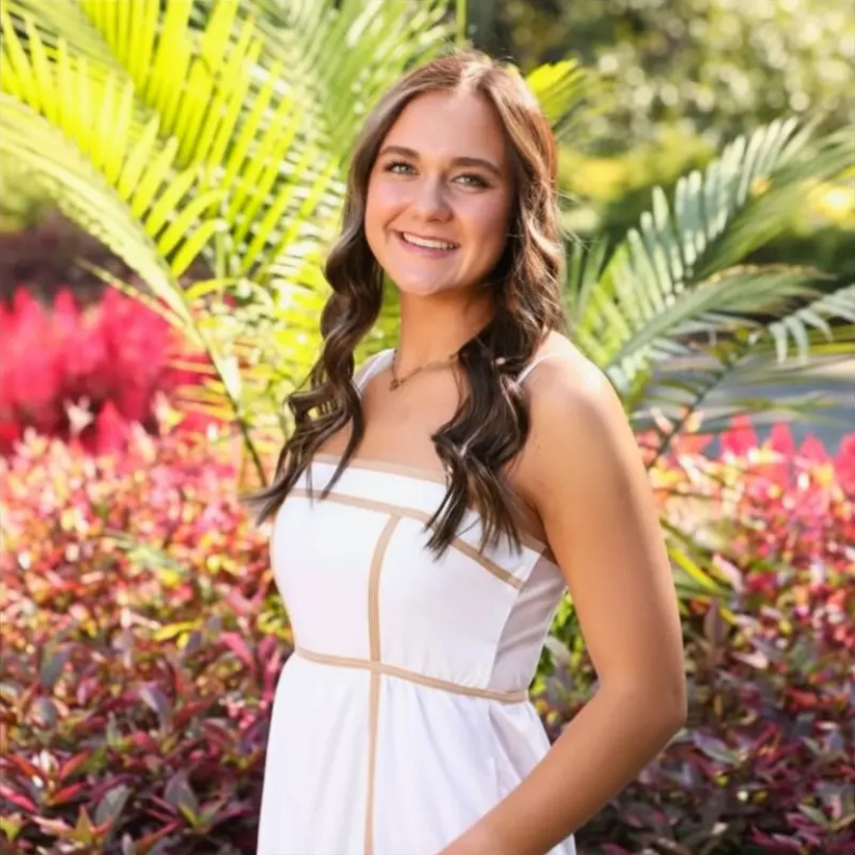 Smiling woman in a white sundress stands in a sunlit tropical garden with lush greenery behind her.