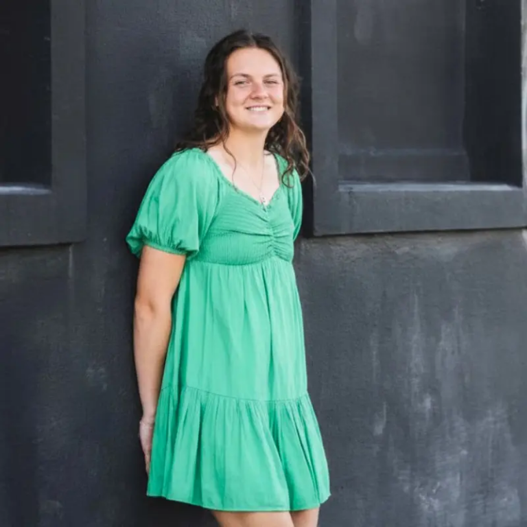 Young woman in a bright green dress standing against a dark wall, smiling at the camera.